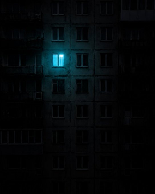 A dark, moody photograph of an apartment building facade at night, with a single brightly illuminated cyan window glowing among the otherwise dark, unlit windows.