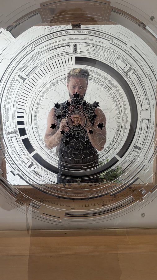 A man holding a Bitcoin-branded medallion stands behind a circular sci-fi-style display, with a Bitcoin emblem visible on his chest.
