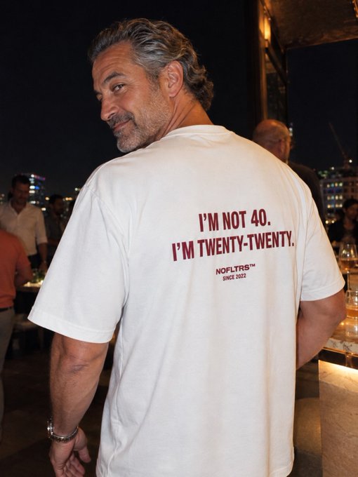 A man with grey hair looks back over his shoulder at the camera, wearing a white t-shirt that reads 'I'M NOT 40. I'M TWENTY-TWENTY. NOFLTRS SINCE 2022'.