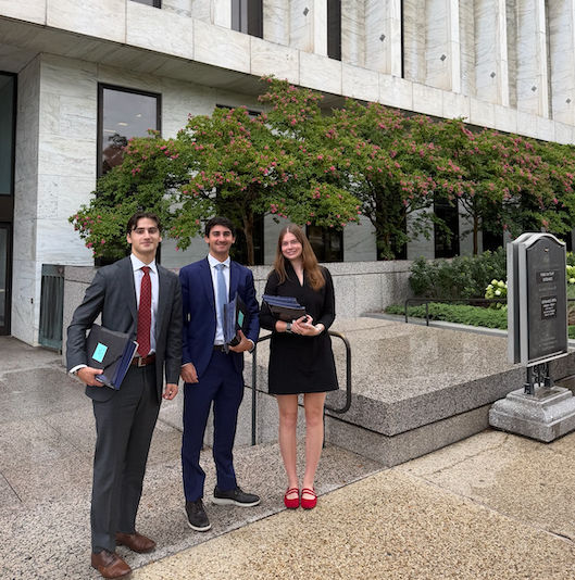 Three professionally dressed people—two men in suits and a woman in a black dress holding books—pose outside a stone building next to a historical marker.