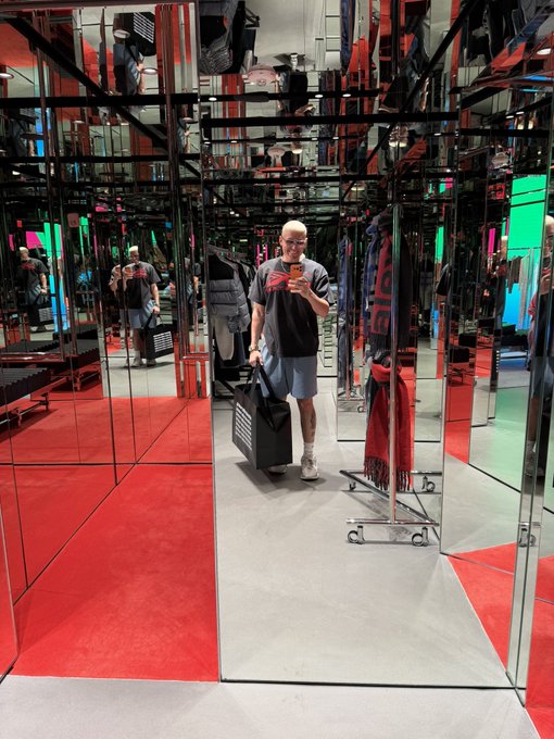 A young man wearing glasses takes a mirror selfie inside a visually striking, mirrored retail space with bright red carpeting, while wearing a graphic t-shirt and holding a shopping bag. The mirror maze-style dressing room reflects him from multiple angles, capturing a stylish weekend shopping moment.
