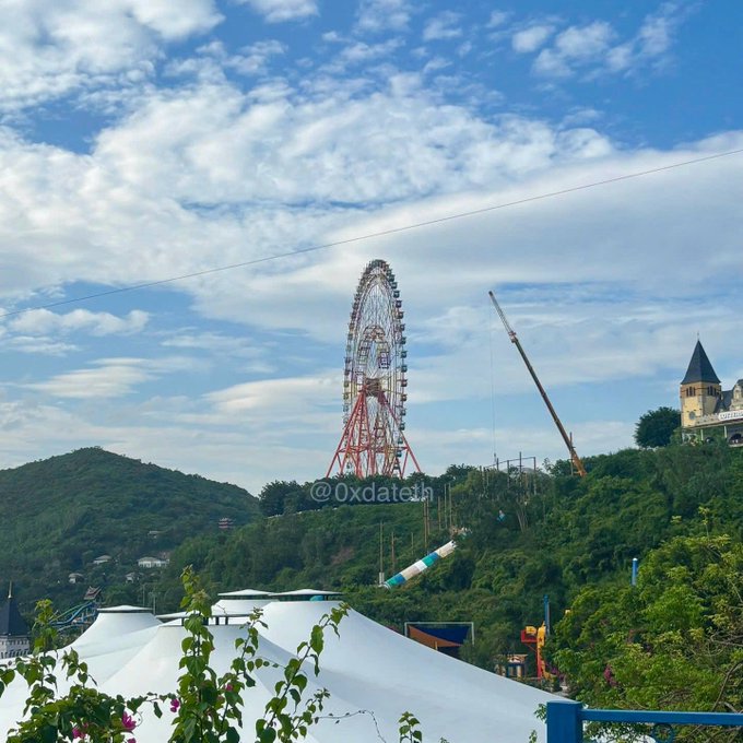 A large Ferris wheel and a castle-like building are visible against a partly cloudy blue sky, with a crane nearby, viewed from behind a white tent and trees, watermarked @0xdateth.