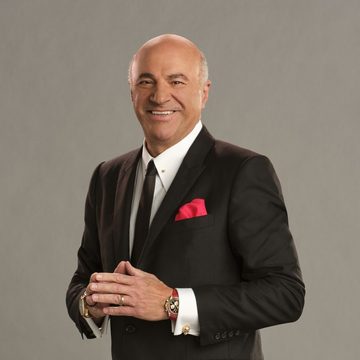 A portrait photograph of a bald man in a dark suit with a pink pocket square and white shirt, smiling against a plain gray background.