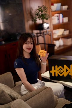 A young woman sits on a couch in what appears to be a Binance office lounge, smiling and holding up a small yellow toy figure, with the partially visible orange "BINANCE" logo displayed on the table beside her—visually connecting to the post celebrating Binance's 9-year journey through major crypto milestones alongside the comment about childhood versus early industry experience.