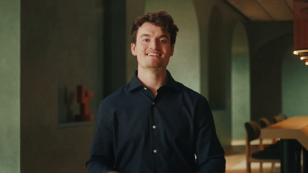 The image features a man wearing a blue shirt standing in front of a dining table with chairs around it. The man appears to be smiling at the camera while posing for the photo. The background includes a green wall, which complements the man's attire. The image also contains text that reads "Claude Fable 5 more than doubles the previous best Claude on the hardest coding benchmark most people will fumble it before the free window even closes". This text seems to be related to the post about Claude Fable and his impressive skills in computer programming.