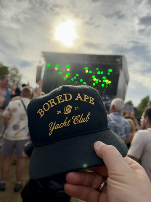A hand holds up a black 'Bored Ape Yacht Club 2021' cap at an outdoor event, with a stage featuring green and yellow lights visible in the sunlit background.