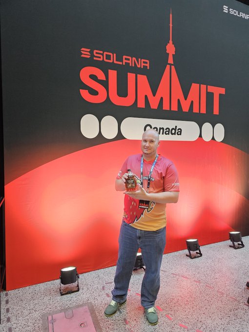 A man wearing a red and yellow flame-patterned t-shirt and blue jeans stands in front of a "SOLANA SUMMIT Canada" backdrop, holding a small device.