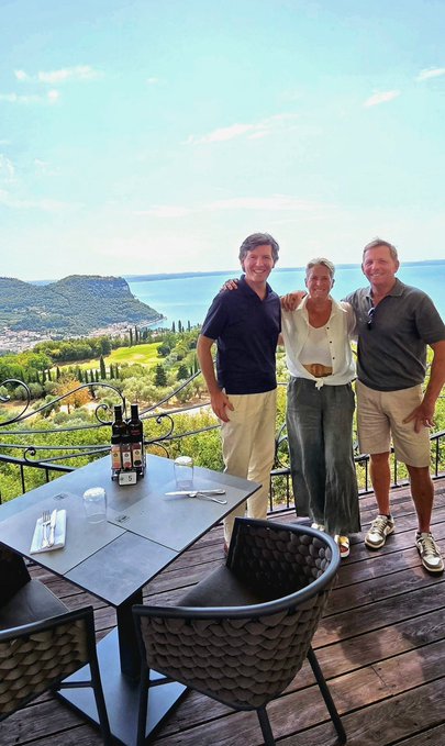 Three people posing together on a scenic outdoor terrace overlooking a coastal landscape with a body of water and mountains in the background, with a table holding wine bottles and glasses.
