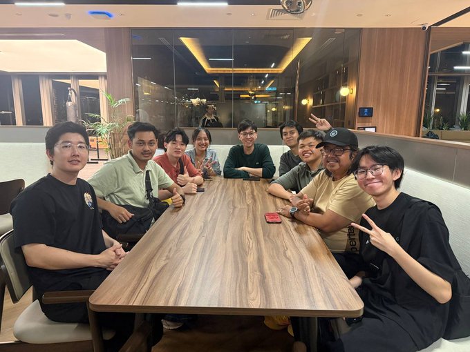 A group of nine people pose together at a wooden conference table, smiling and making peace signs, with several wearing casual clothing including a man in a black t-shirt and a man in a Supreme cap.