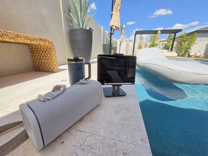 A portable JBL Bluetooth speaker and tablet sit on a sunlit poolside ledge, paired with a navy Stanley tumbler, as the user's "office" for monitoring their successful short positions on Bitcoin. The luxurious backyard setting with lounge chairs and umbrella reflects the "Life is good!" sentiment from cashing in on the BTC trade.