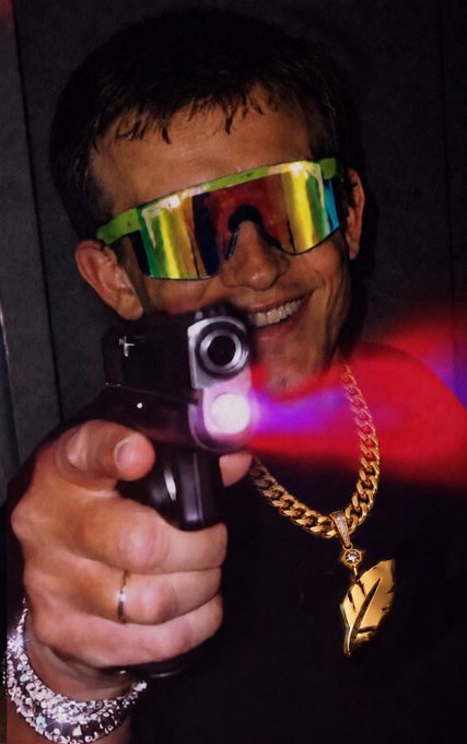 A smiling young man wearing colorful reflective ski goggles, a gold chain with 'LV' pendant, and bracelets points a handgun with a red muzzle flash toward the camera.