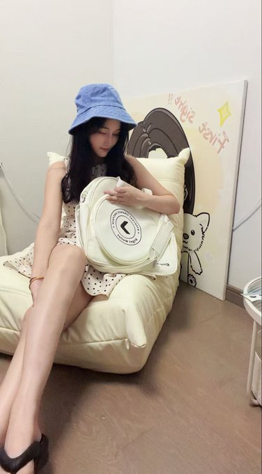A young woman with long dark hair, wearing a blue bucket hat, a polka dot dress, and black heels, sitting on a cream-colored sofa holding a light green backpack, with a canvas reading 'Magice star' propped up nearby.