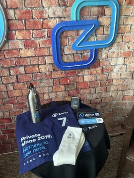A 7th birthday swag bag display for "Zano," featuring a purple "Private Since 2019" t-shirt and water bottle against a brick wall with a blue "Z" logo. The caption references celebrating Zano's 7-year anniversary and the accompanying promotional merchandise.