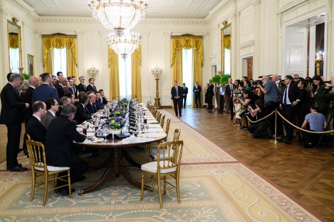 A formal meeting takes place in an ornate room with chandeliers and yellow curtains, where men in suits sit at a long table while a man in a dark suit stands at the head addressing them, with a crowd of photographers documenting the event.
