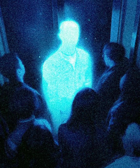 A glowing blue translucent figure of a man in a suit standing in a dark room with people around him.