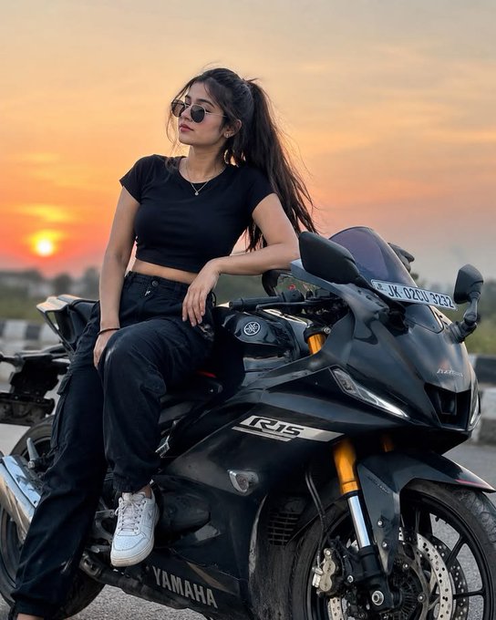 A young woman wearing sunglasses and a black t-shirt sits on a black Yamaha R15 motorcycle during sunset, with the license plate 'JK 02EU 9232' visible.