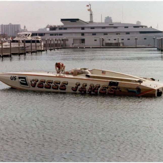 A speedboat named "JESSE JAMES" with "US 3" on its side floating on the water, with a large yacht docked in the background.