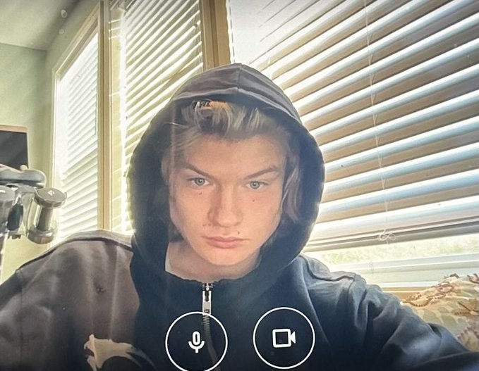 A young man wearing a black hoodie looks directly at the camera with a video call interface showing microphone and camera buttons visible at the bottom of the frame.