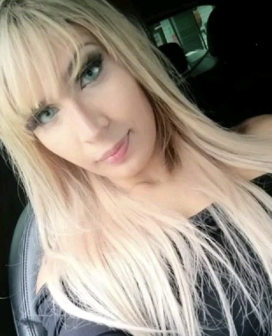 A close-up selfie of a woman with long blonde hair, bangs, and blue eyes sitting inside a vehicle.