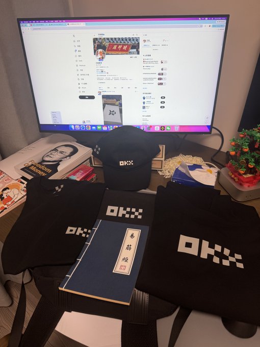 A computer monitor displays a website, with merchandise including black bags, shirts, and a hat featuring the logo 'OKX' displayed in front.