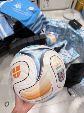 A hand holding a white, blue, and orange soccer ball with patches for "AF" and "Argentina" visible, with other merchandise scattered on a tiled floor in the background.