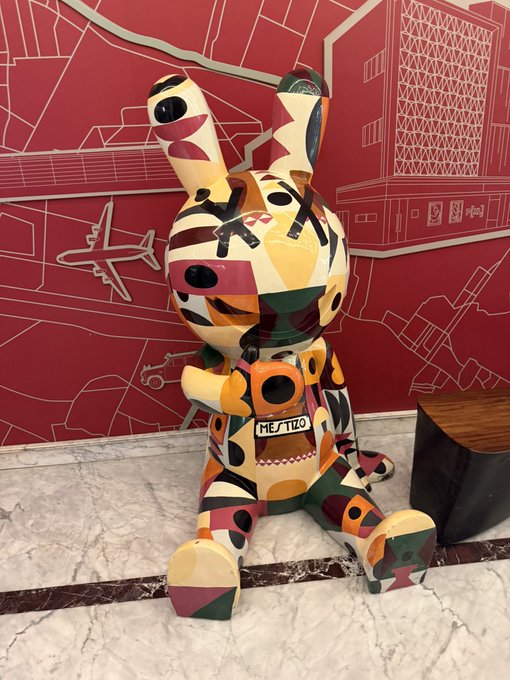 A colorful, geometric-painted rabbit sculpture with a tag reading 'ME/TIZO' sits on a marble floor in front of a red illustrated wall.