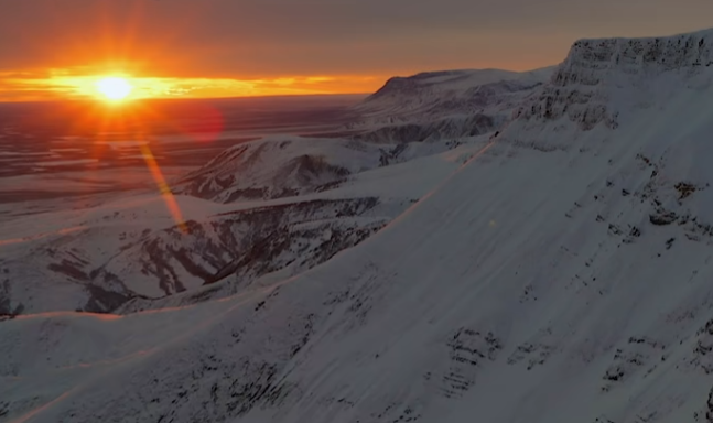 An aerial landscape photograph capturing a bright sunset over a snow-covered mountain range with the sun's rays creating a lens flare across the icy terrain.