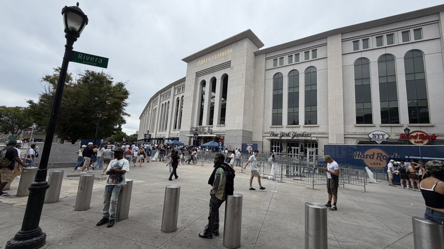 An exterior view of Yankee Stadium with crowds of people gathered outside, featuring a street sign for 'River Ave' and a Hard Rock Cafe visible on the building.