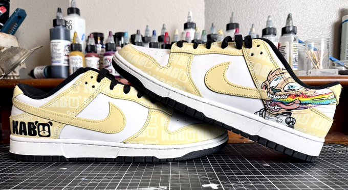 Custom-painted Nike Dunk sneakers in cream and white with 'KABO' text and a cartoon character design, displayed on a workbench surrounded by paint bottles.