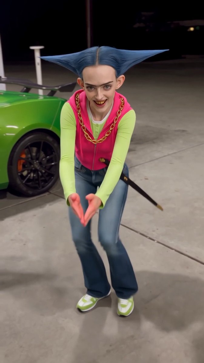 A cosplayer with blue horn-like hair leans toward the camera and forms a heart with both hands beside a green car in a garage.