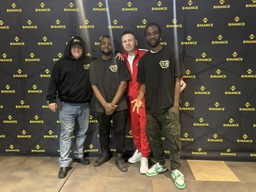 The image features a group of four men posing together against a black backdrop with yellow text, likely representing a company or brand name. The men are standing close to each other, smiling and looking at the camera. One man has his arm around another man's shoulder in a friendly gesture. They all appear to be dressed similarly, wearing matching outfits that include red pants and green shoes.