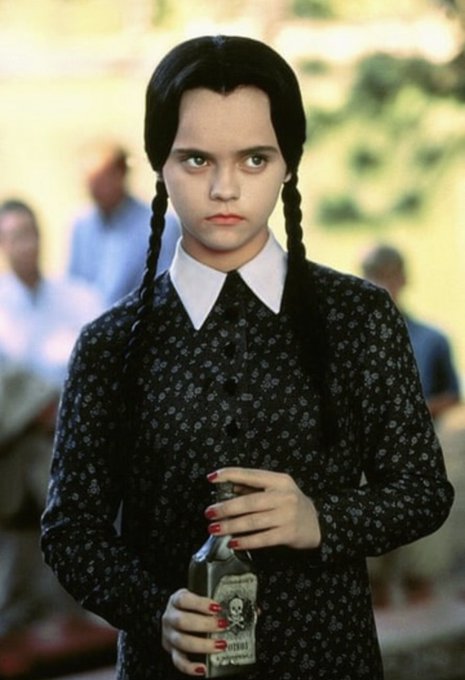 Wednesday Addams, played by Christina Ricci, stands with braided hair wearing a black patterned dress, holding a glass bottle with a skull label.
