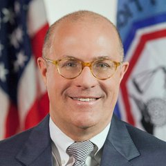 An official portrait of a smiling, bald man wearing glasses, a blue suit, and a gray tie, standing in front of American flags.