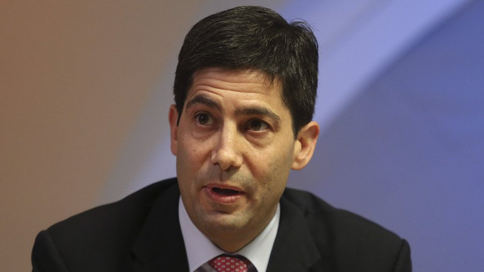 A man in a dark suit with a red patterned tie, appearing to be mid-speech, is shown against a blue and orange background. The image accompanies a statement from Federal Reserve Chair Kevin Warsh regarding the Fed's stance against bailing out crypto firms.