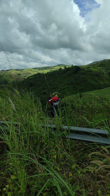 A hiker in a red backpack walking through tall grass along a mountain trail under a cloudy sky.