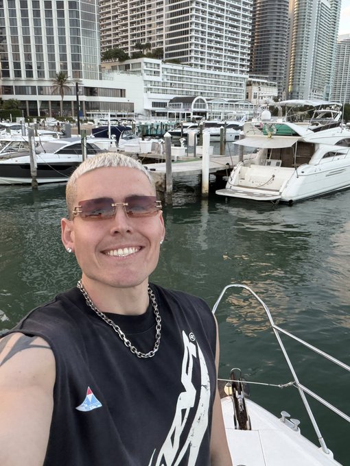 A smiling man with short hair and sunglasses takes a selfie on a boat in a marina, with yachts and high-rise city buildings in the background.