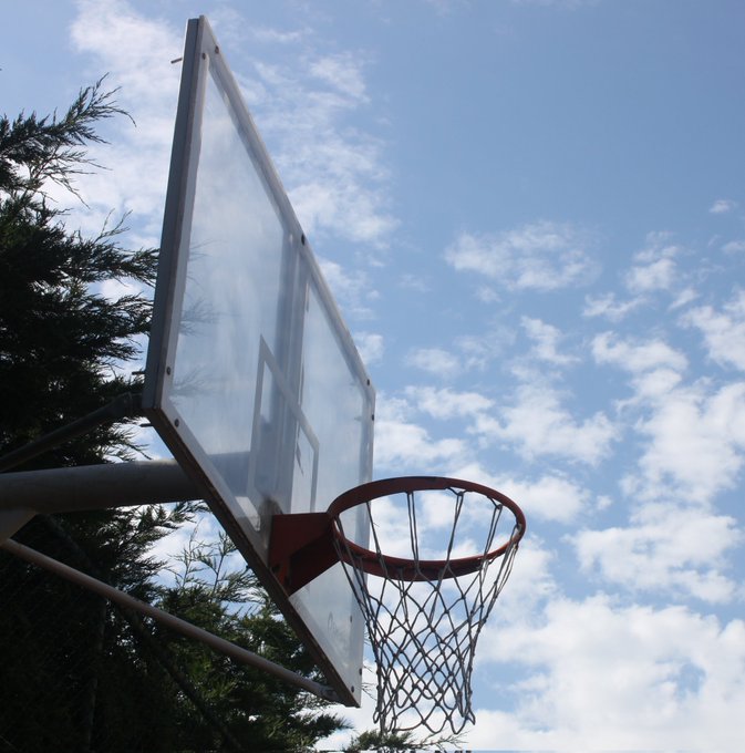 The image features a basketball hoop with a net attached, set against a blue sky with white clouds. The basketball hoop appears to be made of metal or plastic, as indicated by its color and material. The surrounding area includes trees, adding a natural element to the scene. There is also text in the image that reads "gm CT Now say it back! And I'll gib you pancake new day new opportunities let' s get it". This message seems to be related to the basketball hoop or could be part of an advertisement for a sports-related event, product, or service.