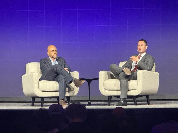 The image shows two men sitting in chairs, engaged in a conversation. One man appears to be wearing a suit, while the other has a tie. They are both seated against a blue wall with white text visible behind them. The background features a large screen displaying information related to cryptocurrency and blockchain technology. This suggests that they might be discussing the latest developments or trends in the crypto world.