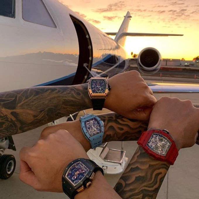 Four tattooed arms display luxury Richard Mille-style watches in different colors (black, blue, red, and black) posed together in front of a private jet at sunset.
