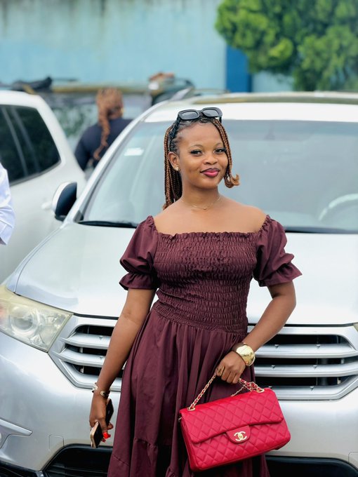 A woman wearing a burgundy off-the-shoulder dress and carrying a red quilted Chanel handbag poses outdoors near a silver vehicle.