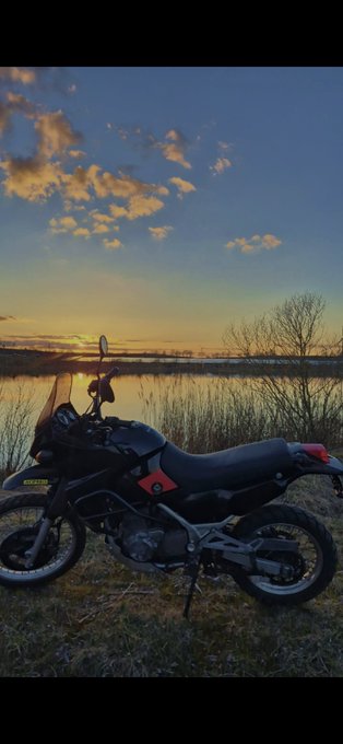A motorcycle is parked on a grassy shore at sunset, with a beautiful sky reflecting on a calm body of water in the background.