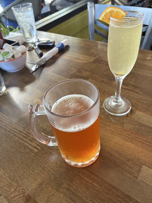 A wooden table set with a glass mug of beer, a champagne flute garnished with an orange slice, and table condiments in the background.