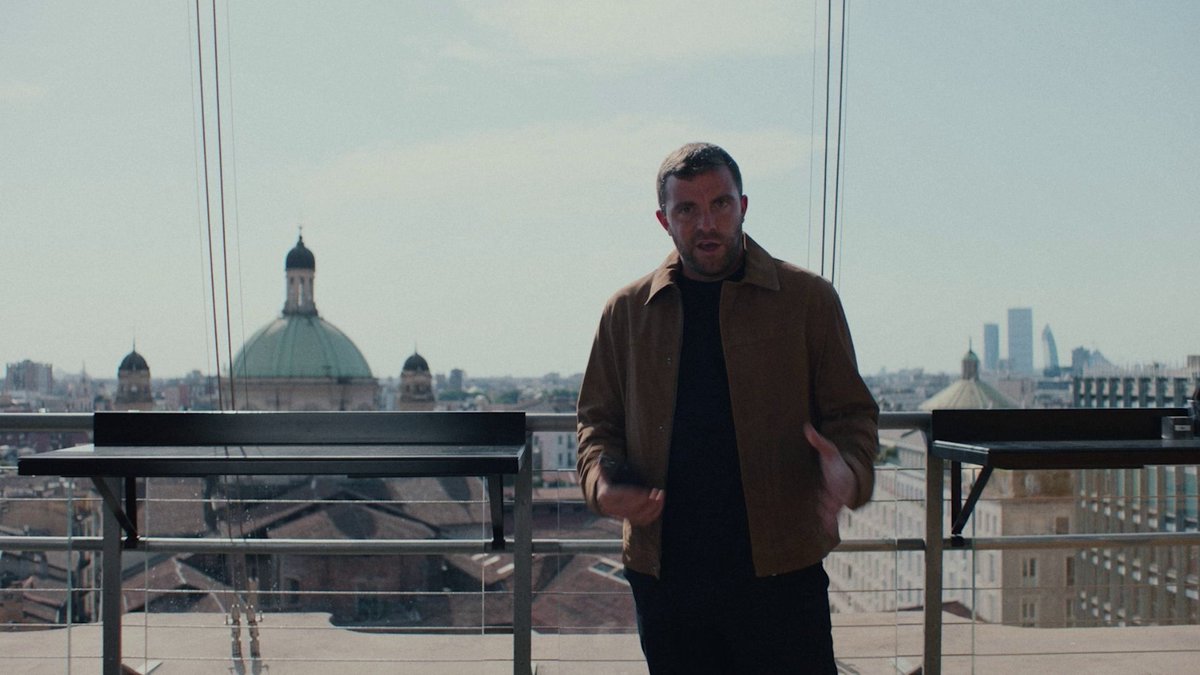 A man in a brown jacket stands on a rooftop balcony overlooking a European cityscape featuring a prominent domed church, gesturing as he speaks into the camera. The cinematic scene likely accompanies a promotional message celebrating a crypto exchange's partnership as an Official Supporter of the FIFA World Cup 2026™.