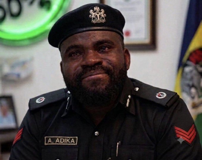A uniformed officer with a name tag reading 'A. ADIKA' smiles, wearing a black beret with a crest and insignia on his shoulders.