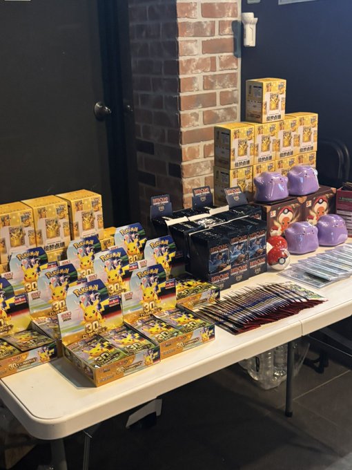 A table displaying various Pokémon merchandise including 30th-anniversary themed boxes, booster packs, and purple Poké Ball-shaped containers, set against a brick wall.