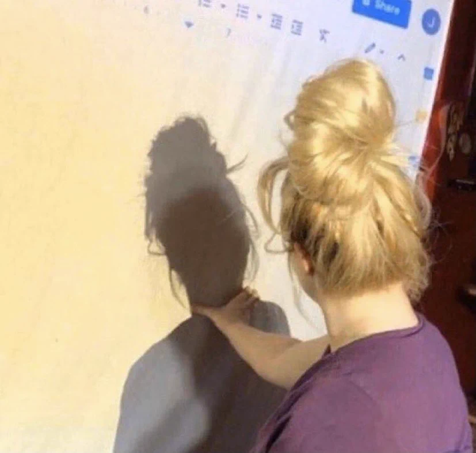A blonde woman with a messy bun stares at a projected screen displaying a Google Doc toolbar with "Share" visible, her shadow cast on the wall beside her—suggesting a tense moment of reviewing documents, humorously paired with the caption lamenting missed early Bitcoin ($BTC) investment.