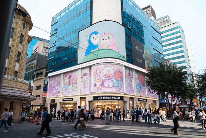 A street view of a multi-story building featuring a large 'LINE FRIENDS SQUARE' storefront, with an LED screen above displaying colorful cartoon characters, surrounded by a busy crowd of pedestrians.