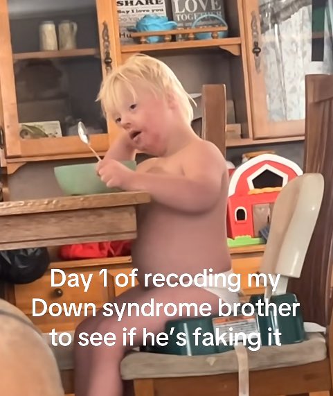 A young child with blond hair sits shirtless in a high chair holding a spoon over a green bowl, with the on-screen text reading "Day 1 of recoding my Down syndrom brother to see if he's faking it." The caption mocks how absurd it is that people on social media now feel the need to "debunk" their own family members, turning genuine disability into viral content for engagement.