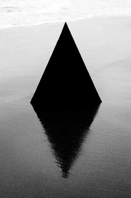 A black and white minimalist image showing a dark triangular pyramid shape reflected in wet sand at a beach.