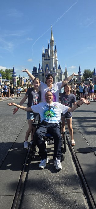 A family poses with arms outstretched in front of Cinderella Castle at what appears to be Walt Disney World, with one family member seated in a wheelchair.